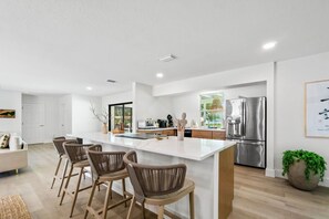 Eine moderne Küche mit einer zentralen Kochinsel, Barhocker, Edelstahlgeräten und einem Essbereich mit einer Couch und Stühlen.