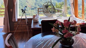 A room with a brick wall, wooden ceiling, a telescope, a hanging chair, a table with a vase of flowers, and a view of a forest. - Chalet 2 - Mountain Range: Design with Hot Tub in the Canyons - Comfort 7km from the Center (Praia Grande)