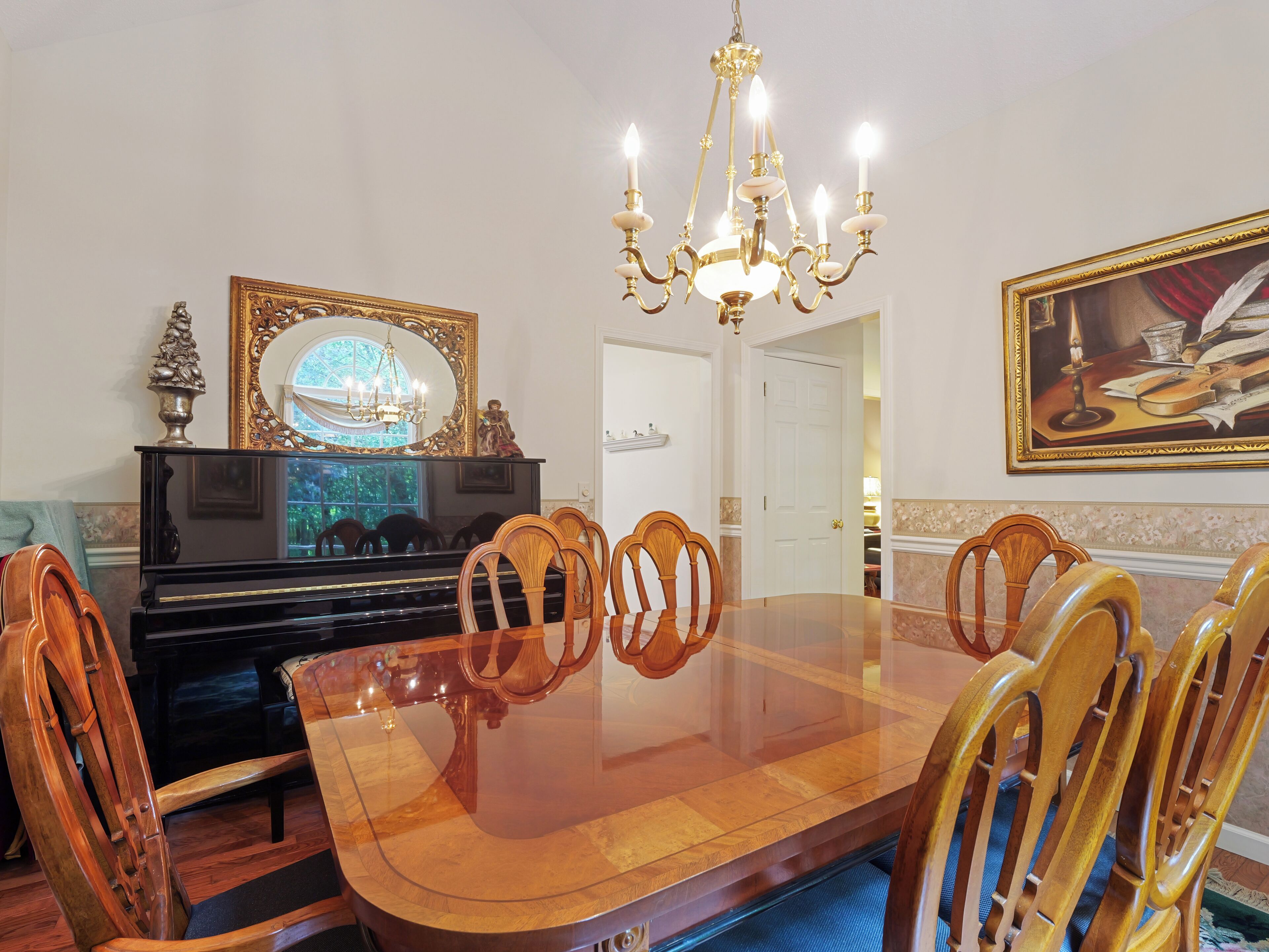 A dining room with a large wooden table, six chairs, a chandelier, a piano, and a painting on the wall.