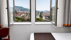A hotel room with a bed, two chairs, and large windows offering a city view.