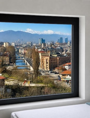 Una habitación con un ventanal grande que da a un paisaje urbano con edificios, un río y montañas.