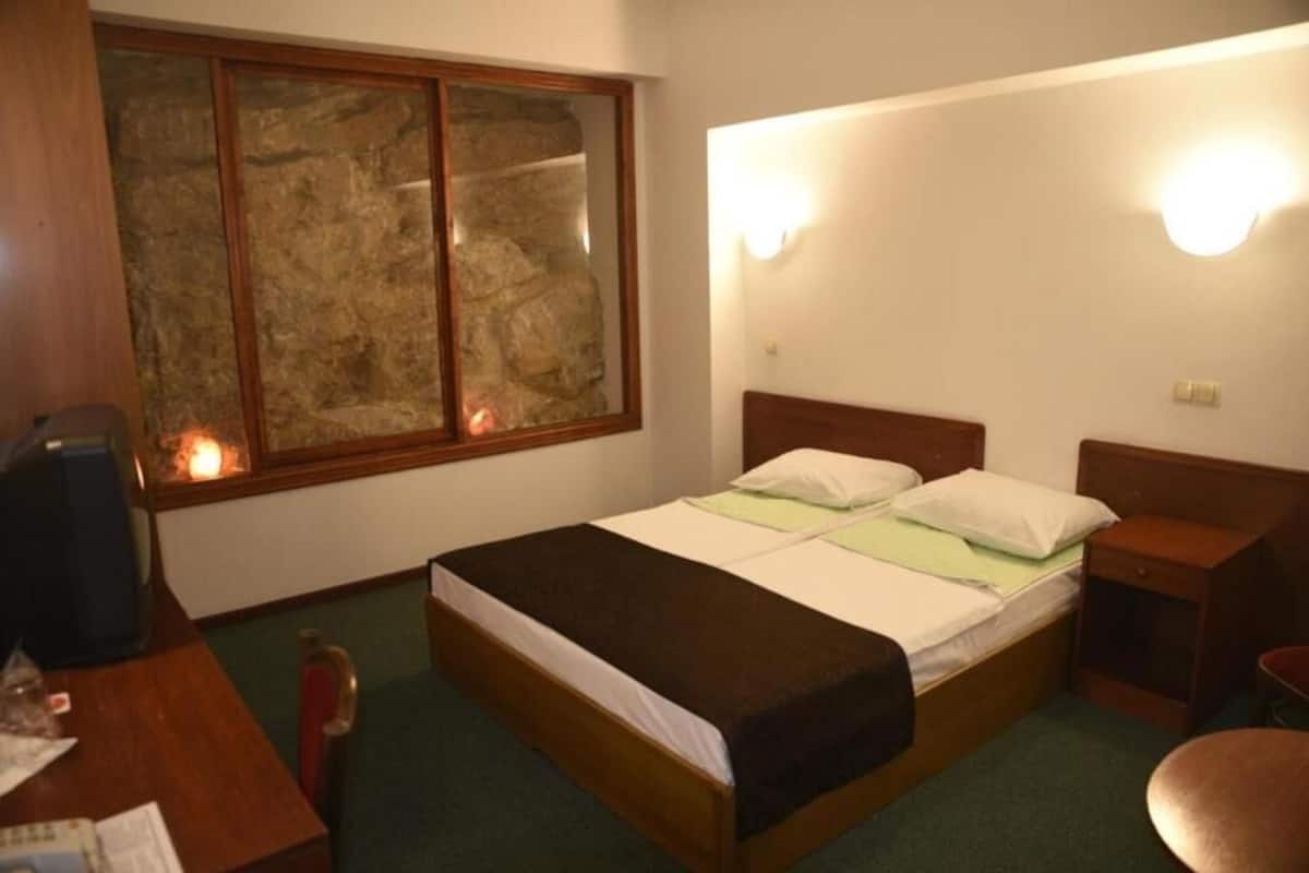 Un dormitorio con cama, mesitas de noche, cabecera de madera, ventana con pared de piedra y lámpara fijada a la pared.