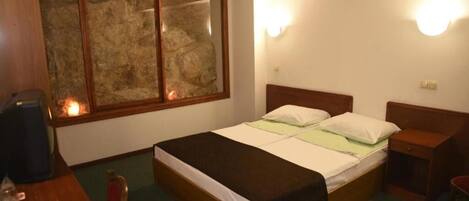 Quarto com cama, mesas de cabeceira, cabeceira de madeira, janela com parede de pedra e luminária fixada na parede.