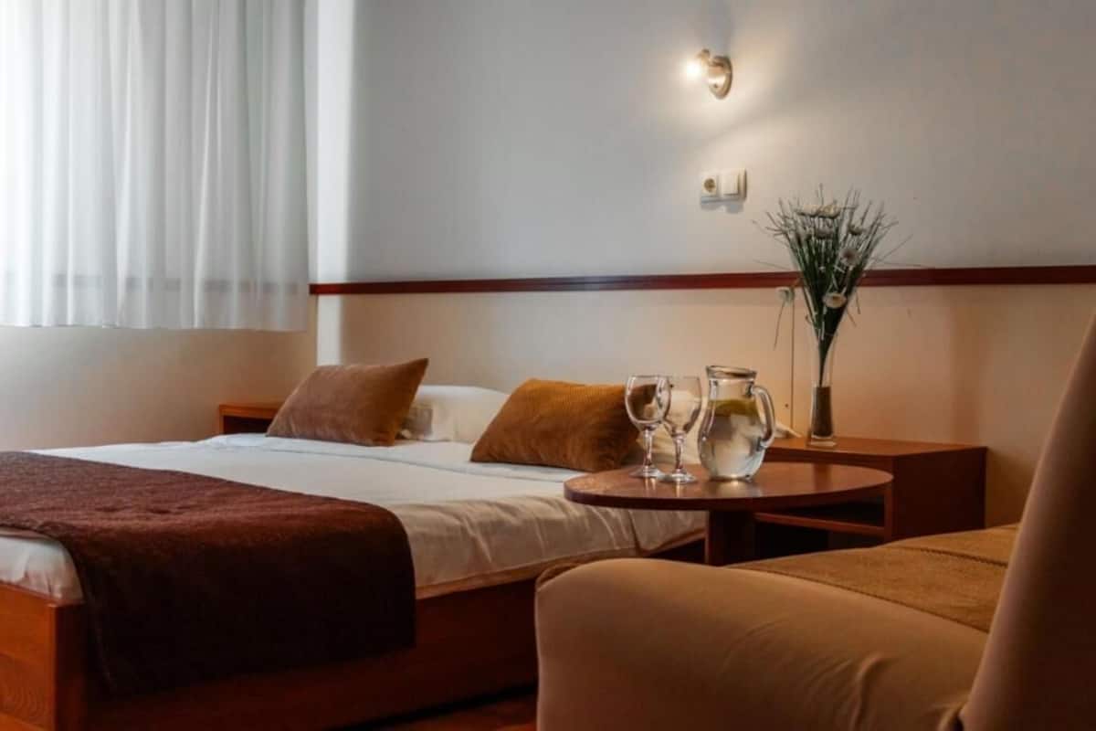 Habitación de hotel con cama, dos almohadas, mesita de noche con un florero y vasos, y ventana con cortinas.