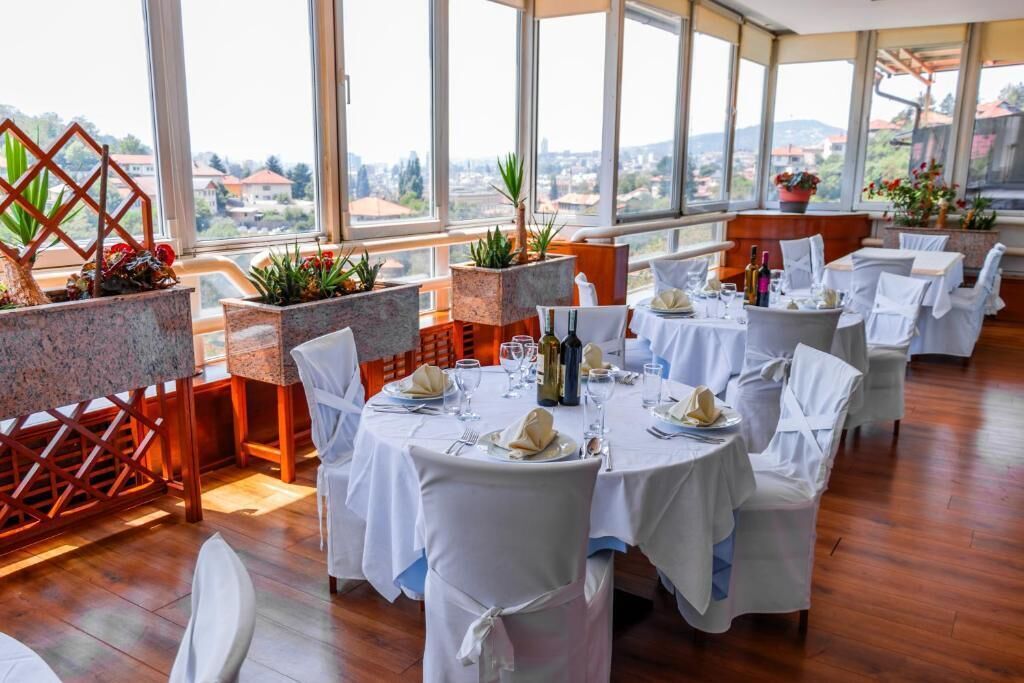 A restaurant with round tables, white tablecloths, and chairs, set against a backdrop of large windows overlooking a cityscape.