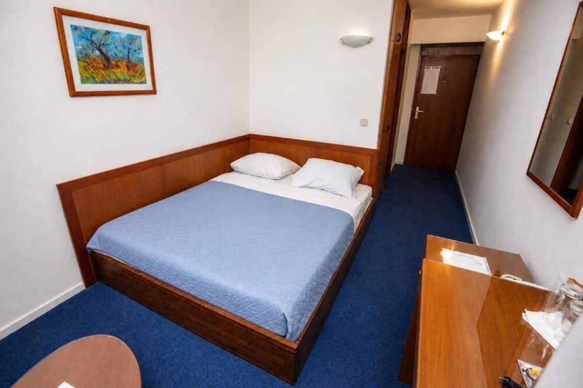 Una cama individual con cabecera de madera, colcha azul y almohadas blancas. Hay un escritorio de madera pequeño con un espejo y un cuadro en la pared.