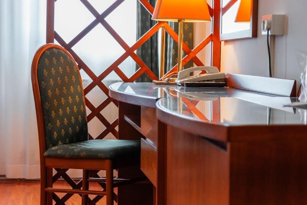 Quarto de hotel com uma cadeira de madeira, uma escrivaninha com abajur e um telefone.