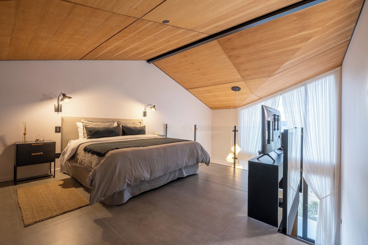 Un dormitorio moderno con techo inclinado, una cama con cabecera, lámparas de noche, una mesita de noche y un ventanal grande con cortinas.