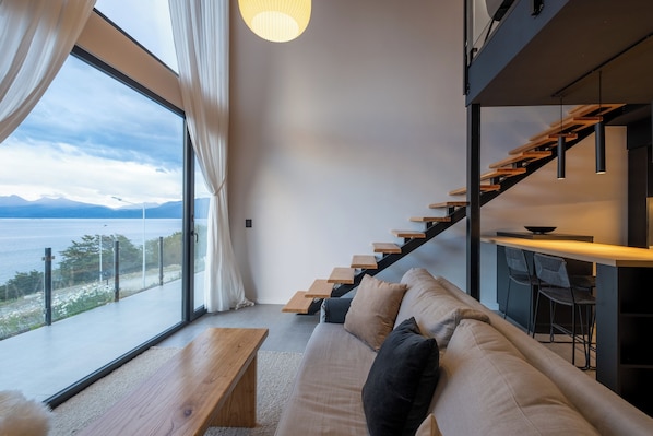 A modern interior with a sofa, wooden staircase, and a view of the sea.