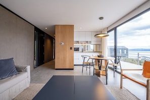 A modern living room with a sofa, a dining table, and chairs, a kitchen area, and a large window with a view.