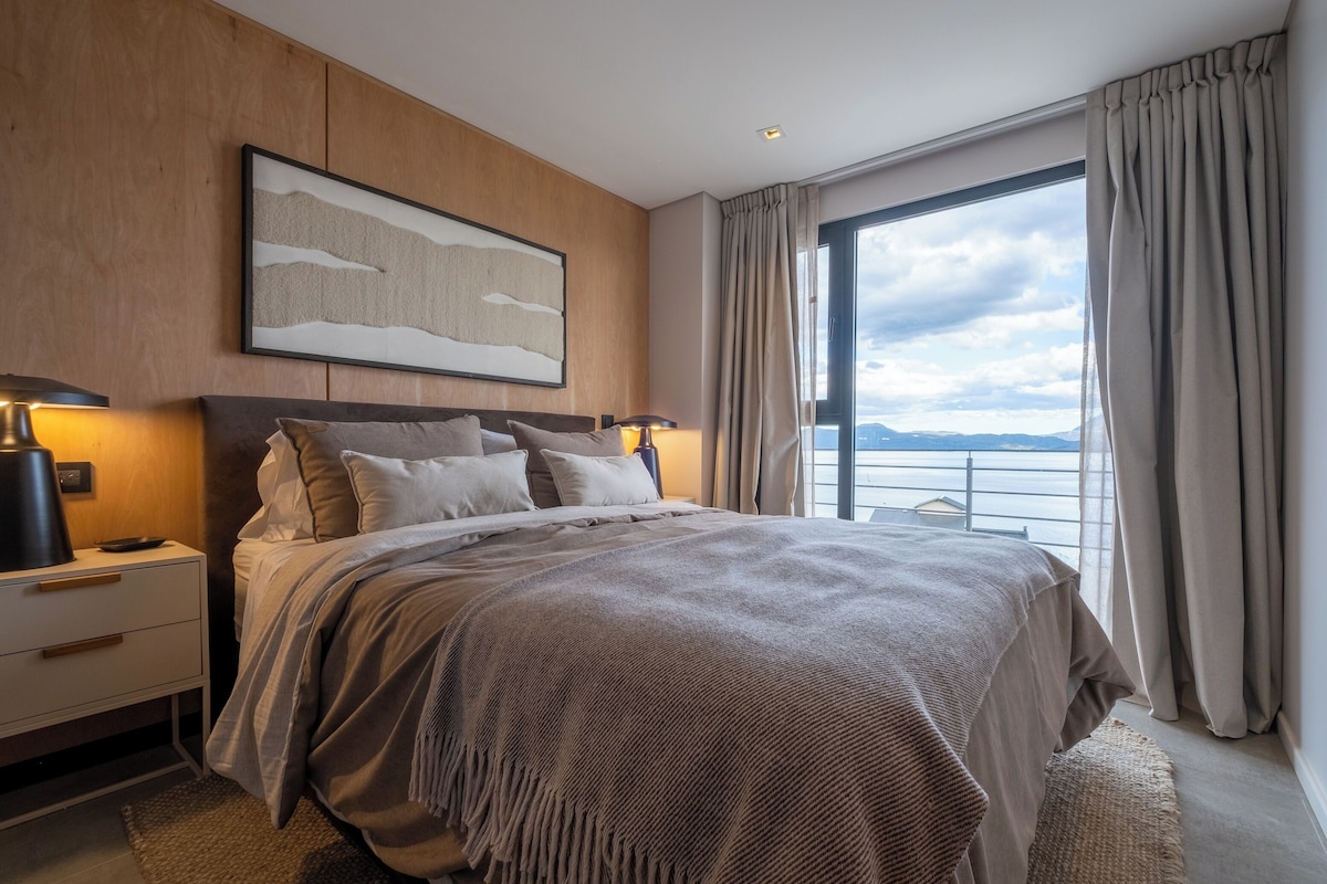 Habitación de hotel moderna con una cama grande, una mesita de noche, una lámpara, una obra de arte enmarcada y vistas al mar.