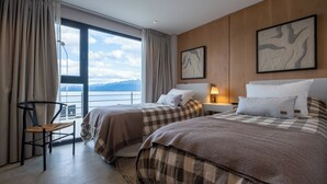 A hotel room with two beds, a chair, a desk, a lamp, and a large window with a view of mountains.