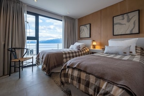 A hotel room with two beds, a chair, a desk, a lamp, and a large window with a view of mountains.
