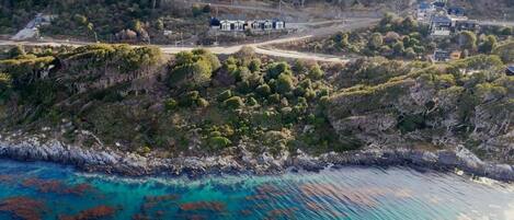 Vista aérea de una propiedad costera con una carretera que conduce a un grupo de edificios, rodeados de vegetación densa y un mar azul cristalino.