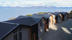 A row of modern, single-story buildings with sloped roofs, positioned on a gravel path with a view of a lake and mountains in the background.