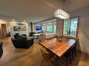 A spacious living area with a wooden dining table, comfortable seating, a fireplace, and a large window providing natural light. - Spacious Family Retreat Near Cowichan Bay. Sleeps 8. Yard, BBQ, Quiet & Private (Cowichan Bay)