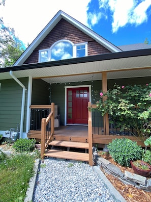 A house with a red door, a porch with a wooden railing, and a garden with various plants. - Spacious Family Retreat Near Cowichan Bay. Sleeps 8. Yard, BBQ, Quiet & Private (Cowichan Bay)