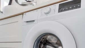 Vue de face d’un lave-linge moderne, de couleur blanche, doté d’un panneau de commande, situé dans un espace cuisine avec un plancher en bois et des meubles blancs.