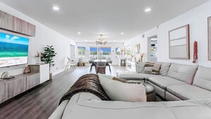 A modern living room with a large sofa, a TV, and a dining area with a view. - Waterfront Villa | BBQ, Pool, Cinema and Tiki (Palm Beach Gardens)
