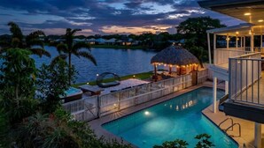 A swimming pool with a thatched-roof structure, surrounded by a fence and palm trees. - Waterfront Villa | BBQ, Pool, Cinema and Tiki (Palm Beach Gardens)