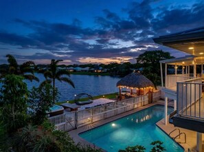 A swimming pool with a thatched-roof structure, surrounded by a fence and palm trees.
