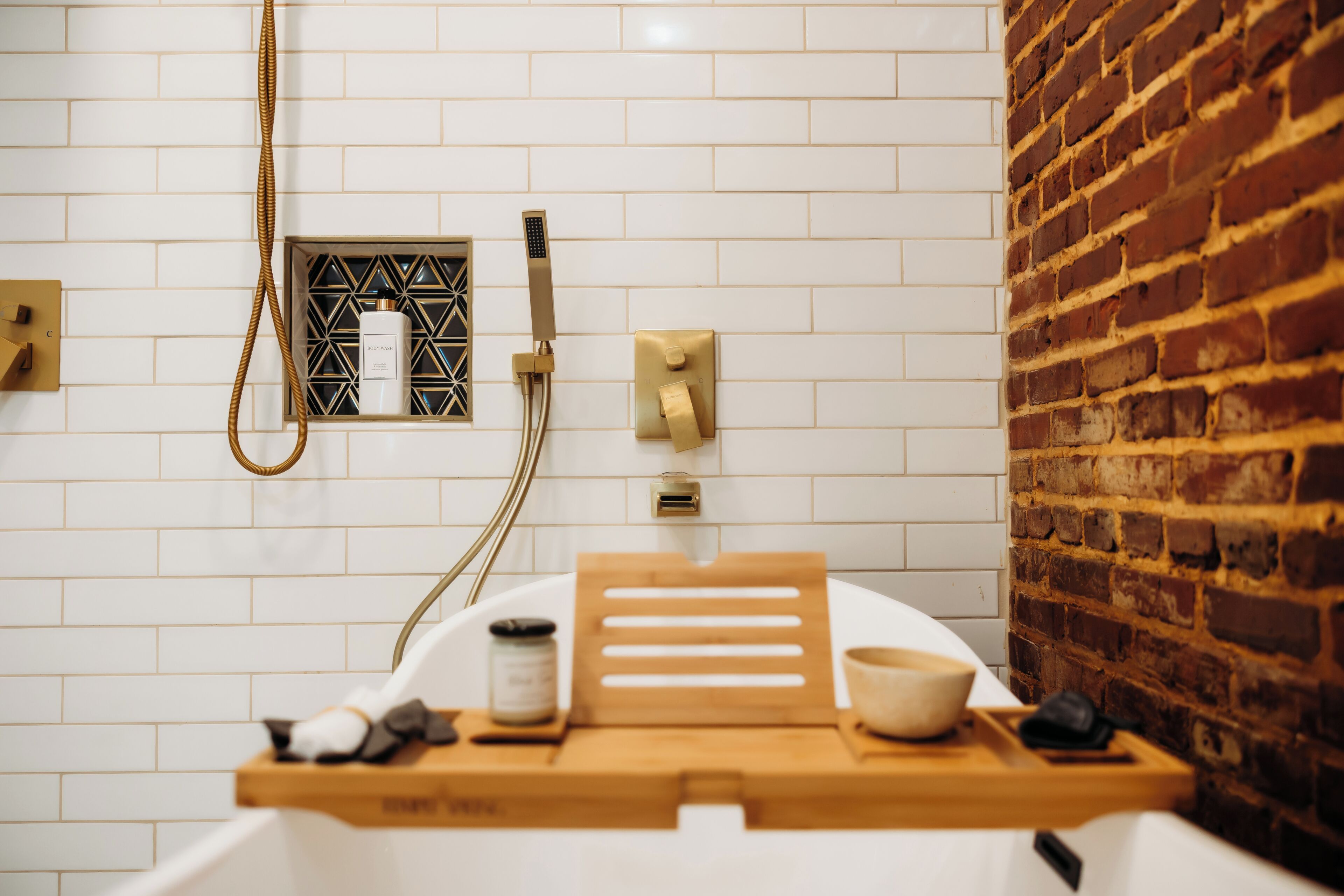 Un baño con paredes de azulejos blancos, una pared de detalles en ladrillo, una ducha con cabezal de ducha manual, un perchero y una bandeja de madera con artículos de aseo.