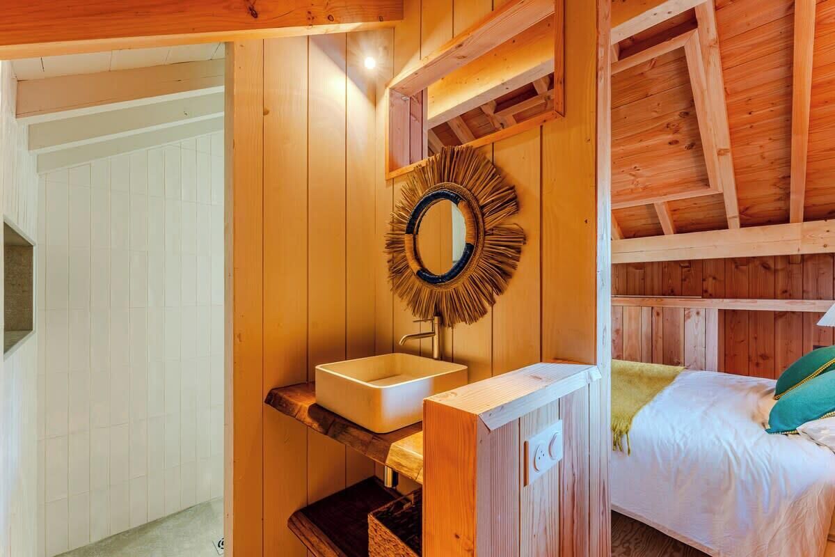 Interior de una cabaña de madera con una cama con estructura de madera, un lavamanos blanco y un espejo con un marco con diseño de sol.