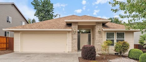 Une maison de plain-pied avec un garage, une porte d’entrée et un petit jardin à l’avant, agrémenté d’aménagements paysagers.