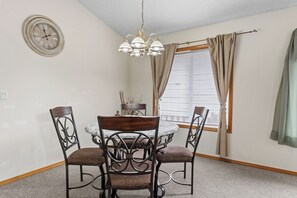 Un comedor con una mesa redonda, cuatro sillas, un reloj de pared y una ventana con cortinas.
