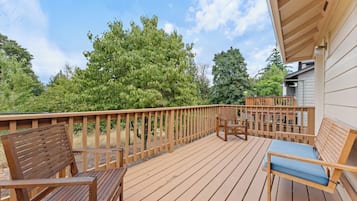 A wooden deck with outdoor furniture, including two wooden chairs and a bench, overlooking a garden with trees and a house in the background.