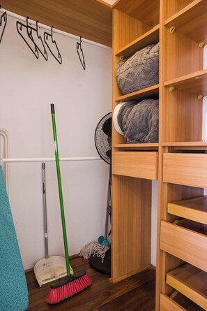 Ein Zimmer mit Besen, Wischmopp und Ventilator auf dem Boden, einem Regal mit gefalteten Decken und einer Wand mit einer einfachen Linienzeichnung.