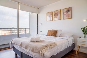 Ein modernes Schlafzimmer mit einem großen Bett, Blick auf die Stadt durch einen Balkon und drei gerahmte Kunstwerke an der Wand.