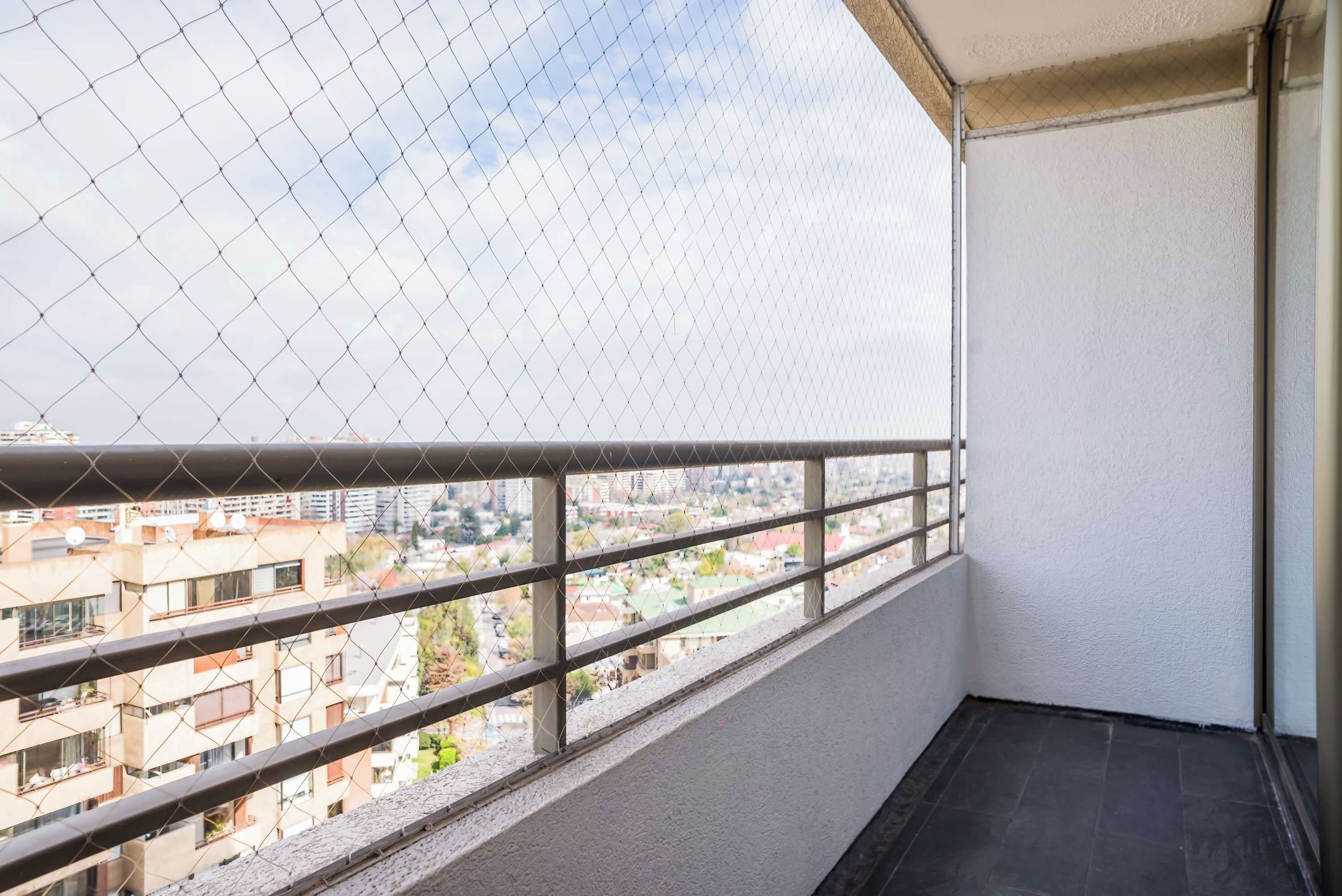 Un balcón con barandilla de metal, vista a un paisaje urbano y una pared blanca.