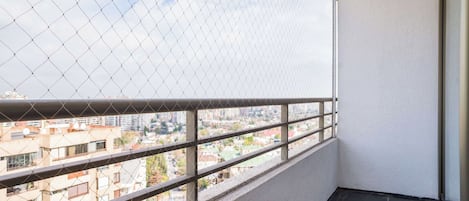Un balcón con barandilla de metal, vista a un paisaje urbano y una pared blanca.