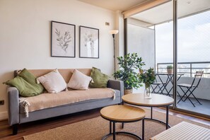 A living room with a grey sofa, two framed botanical prints, a small round table, a potted plant, and a view of a balcony with outdoor furniture. - Chic 2 Bedroom Apartment for 3 with Pool And Gym (Las Condes)