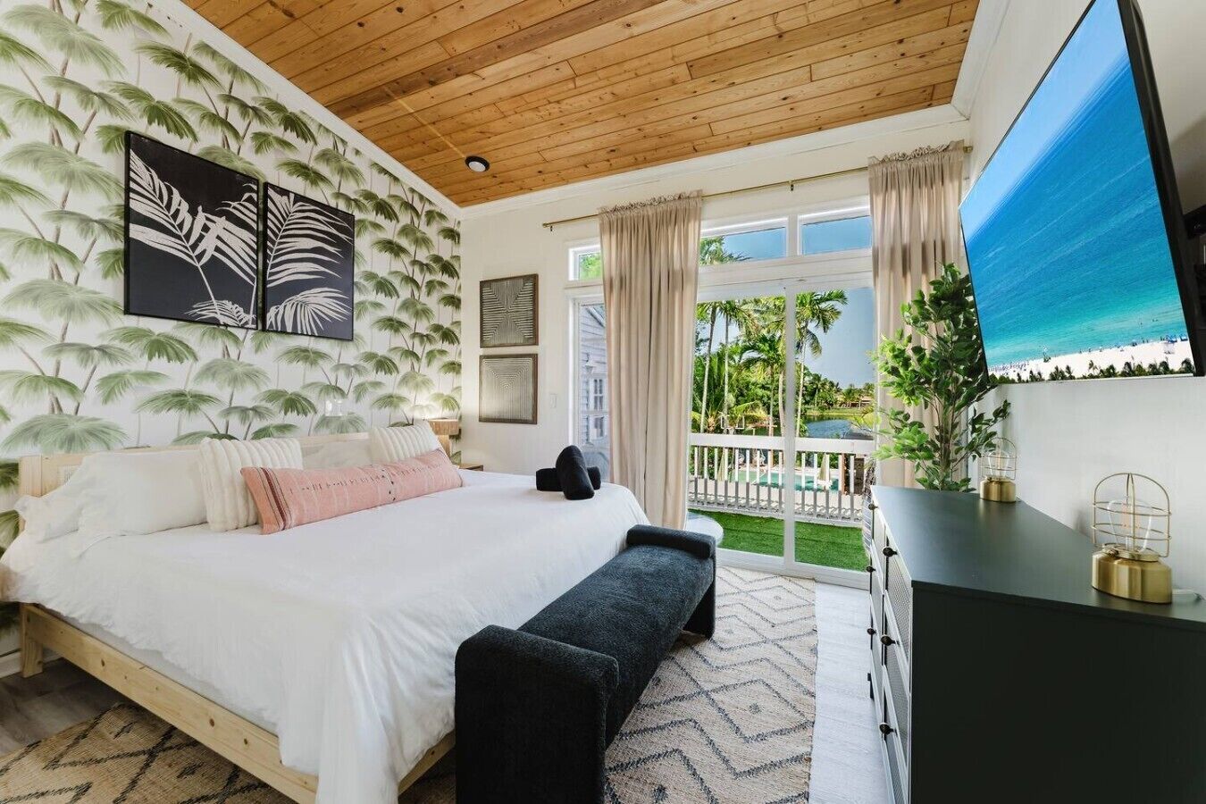 Un dormitorio con cama, un banco, televisión y balcón con vista a la piscina.