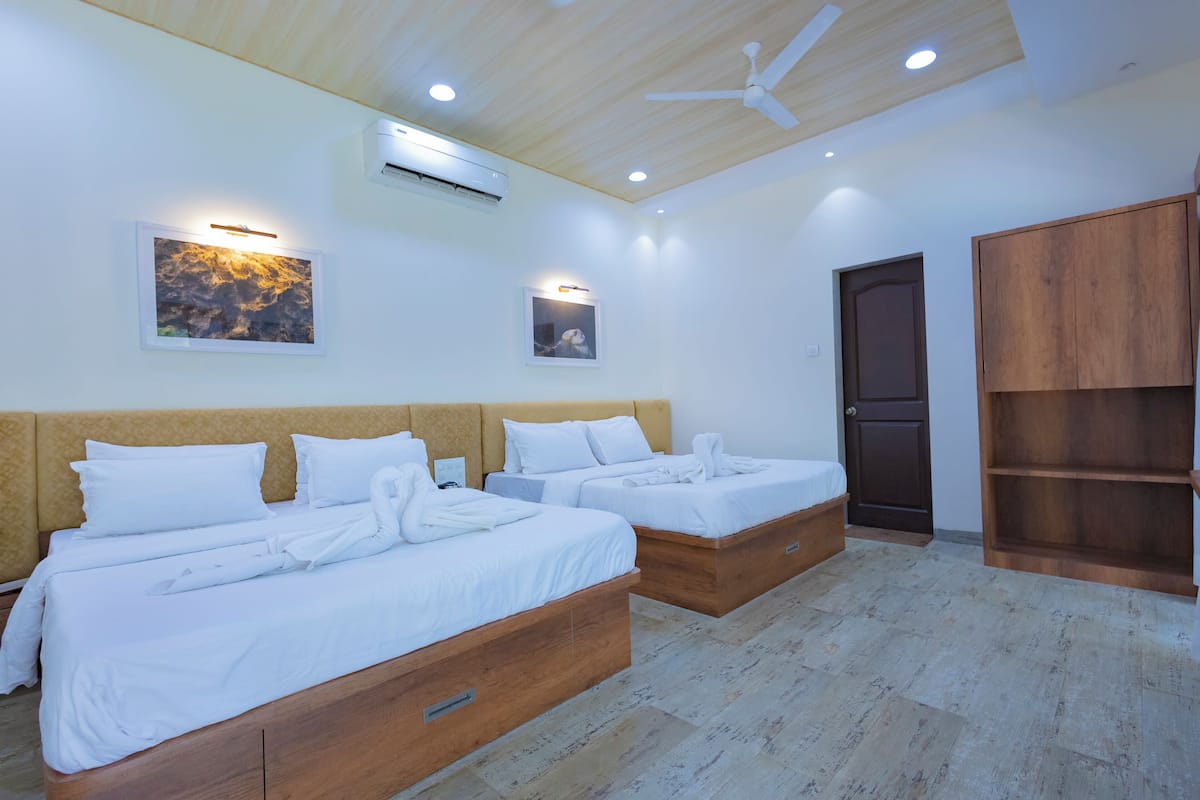 Quarto de hotel com duas camas, cabeceiras de madeira, roupa de cama branca, ventoinha no teto, iluminação embutida e um guarda-roupa de madeira.