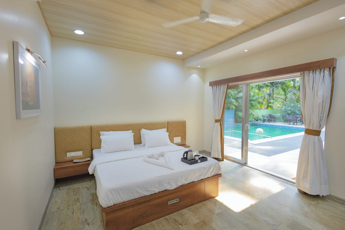 Quarto com cama, mesa de cabeceira, janela com cortinas, ventilador de teto e vista para uma piscina.