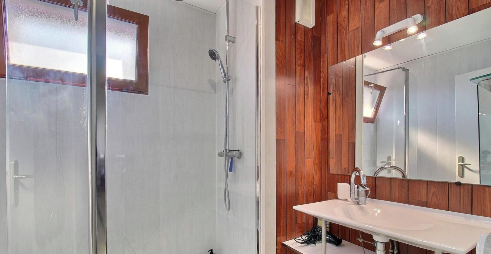 Un baño con cabina de ducha de vidrio, panel de pared de madera, lavamanos blanco y espejo grande.