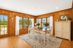 A dining area with wooden paneled walls, a wooden table, four chairs, a cabinet, and large windows with curtains. - Beach front Cottage #3 in Lexington with AC, shared Hot Tub & Pool on Lake Huron (Lexington)