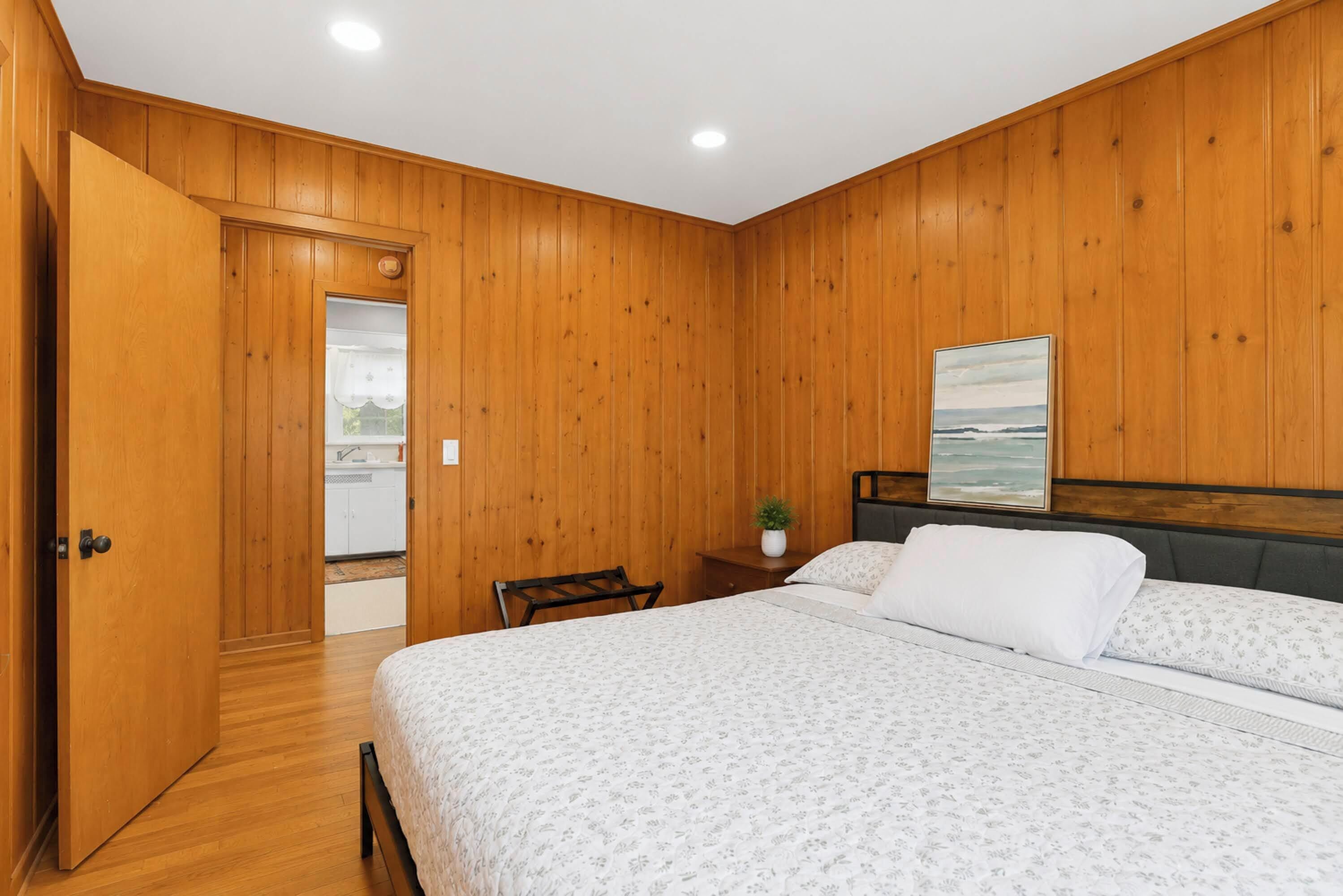 Een slaapkamer met houten lambrisering, een bed met bloemenbeddengoed, een nachtkastje met een plant en een schilderij van een zandstrand.