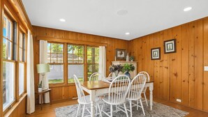 Un comedor con una mesa de madera, sillas blancas, un tapete y una ventana con cortinas.
