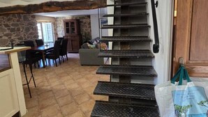 Una escalera con barandilla metálica que lleva a una plataforma, un área de comedor con sillas y una mesa, y una cocina con pared de piedra y una ventana.