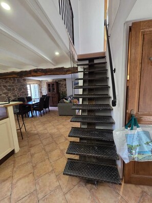 A staircase with a metal handrail leading to a landing, a dining area with chairs and a table, and a kitchen area with a stone wall and a window. - Typical Corsican house (Appietto)