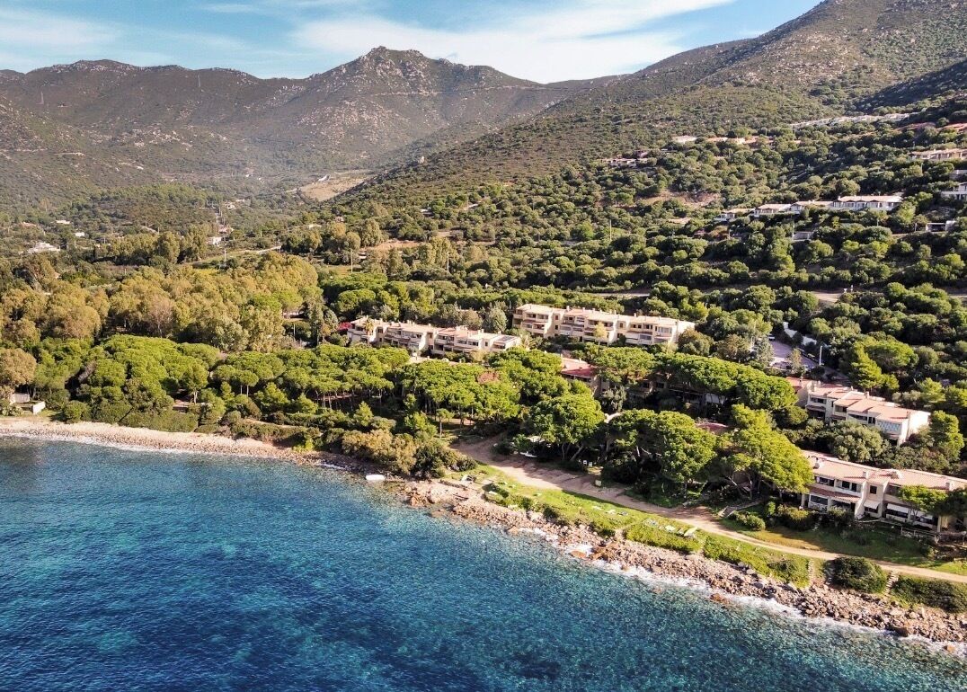 A coastal area with buildings surrounded by dense greenery and mountains in the background.