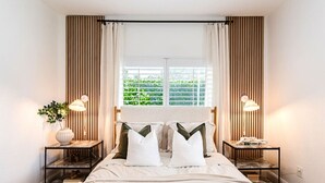 A bedroom with a bed, two bedside tables with lamps, a window with curtains, and a view of greenery outside.