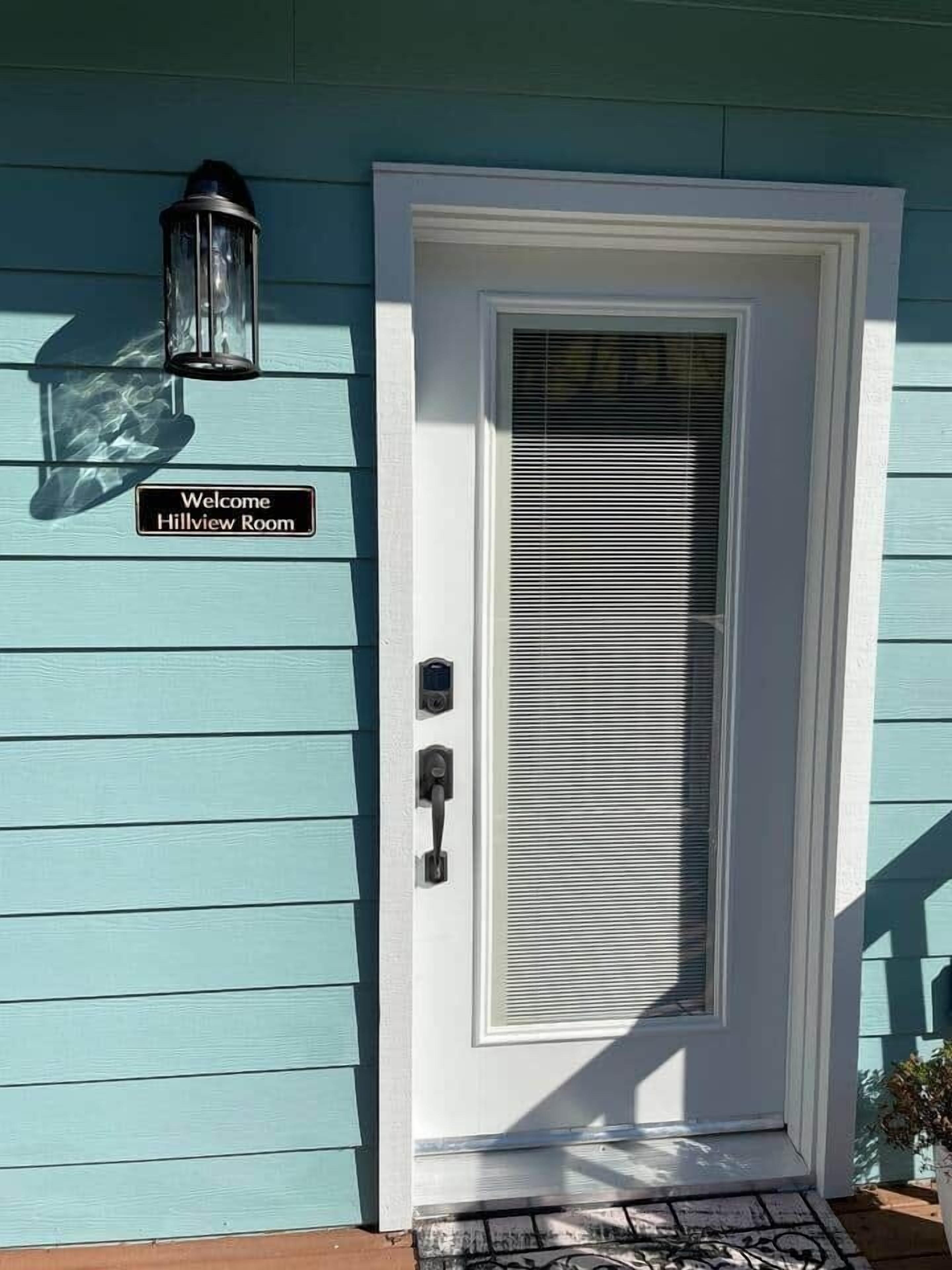 Una casa azul verdoso con una puerta blanca, un cartel que dice 'Welcome Hillview Room', un timbre y una lámpara fijada a la pared.