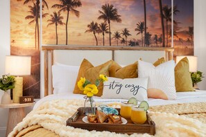 Ein Bett mit Blick auf einen tropischen Sonnenuntergang, ein Tablett mit Toast und Saft sowie ein Kissen mit der Aufschrift 'stay cozy'.