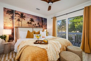 Ein Schlafzimmer mit einem großen Bett, einem Nachttisch mit Lampe, einem Palmen-Gemälde und Blick auf Grün durch das Fenster.