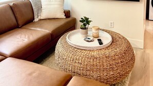 Un salón con un sofá de cuero marrón, un otoman tejido redondo, un televisor de pantalla plana montado en la pared y una planta pequeña sobre el sofá.